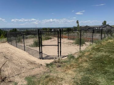 Black chain-link fence with gate encloses a dirt area on a hillside, overlooking a distant cityscape under a blue sky.
