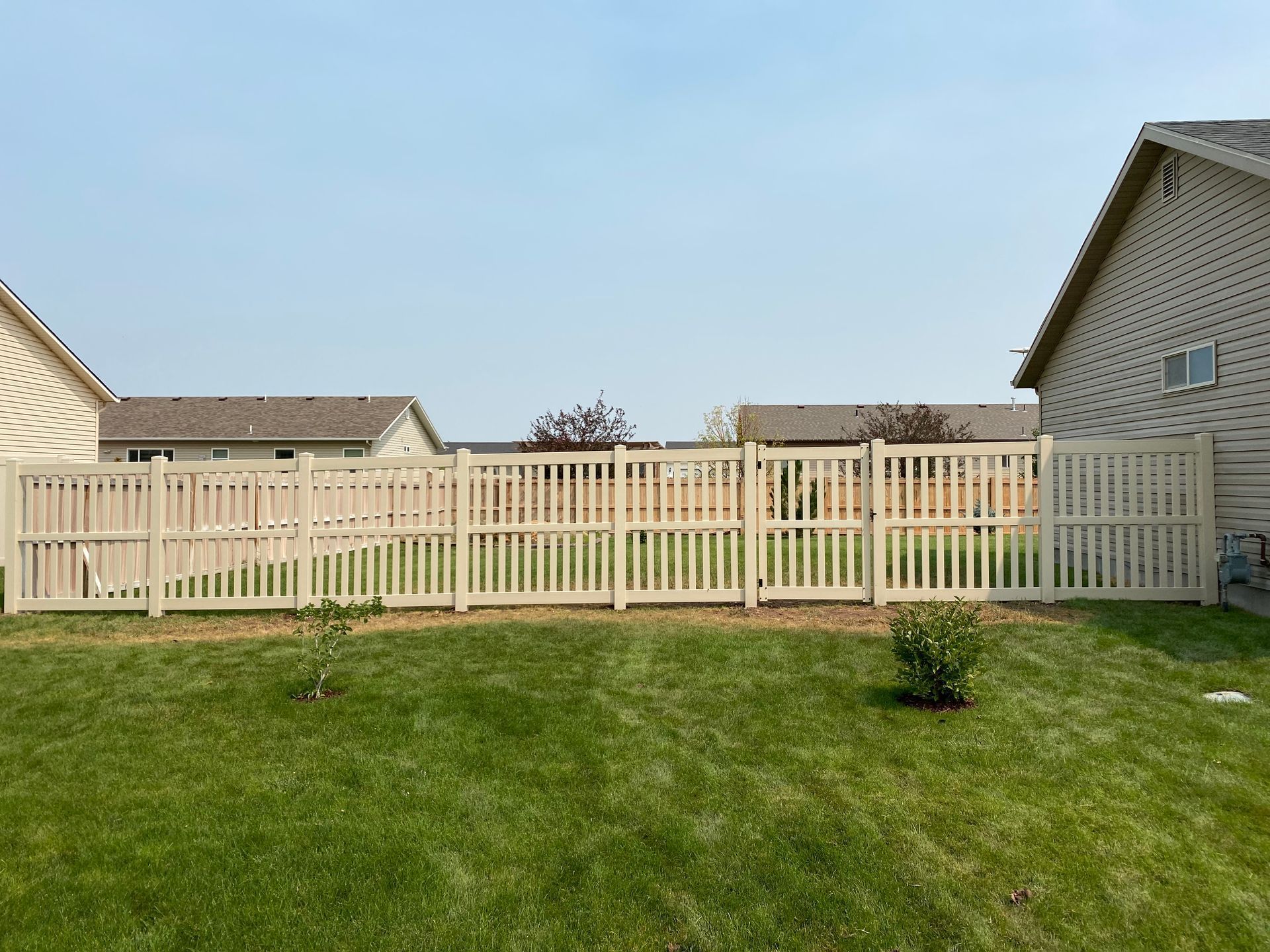Tan picket fence in a green yard, houses in background under a hazy blue sky.