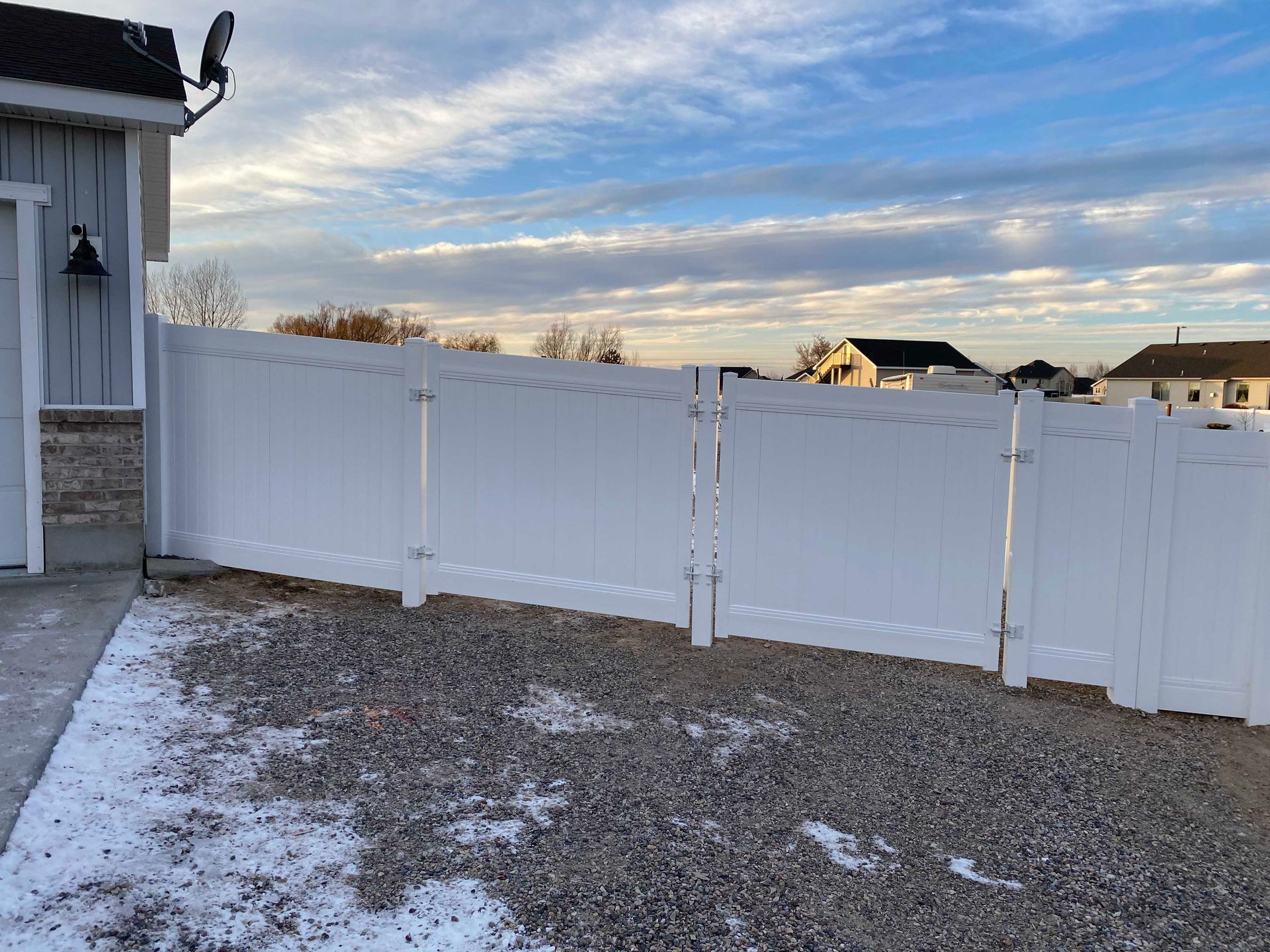 White vinyl fence with double gates in a gravel yard, partly snowy, under a cloudy sky.