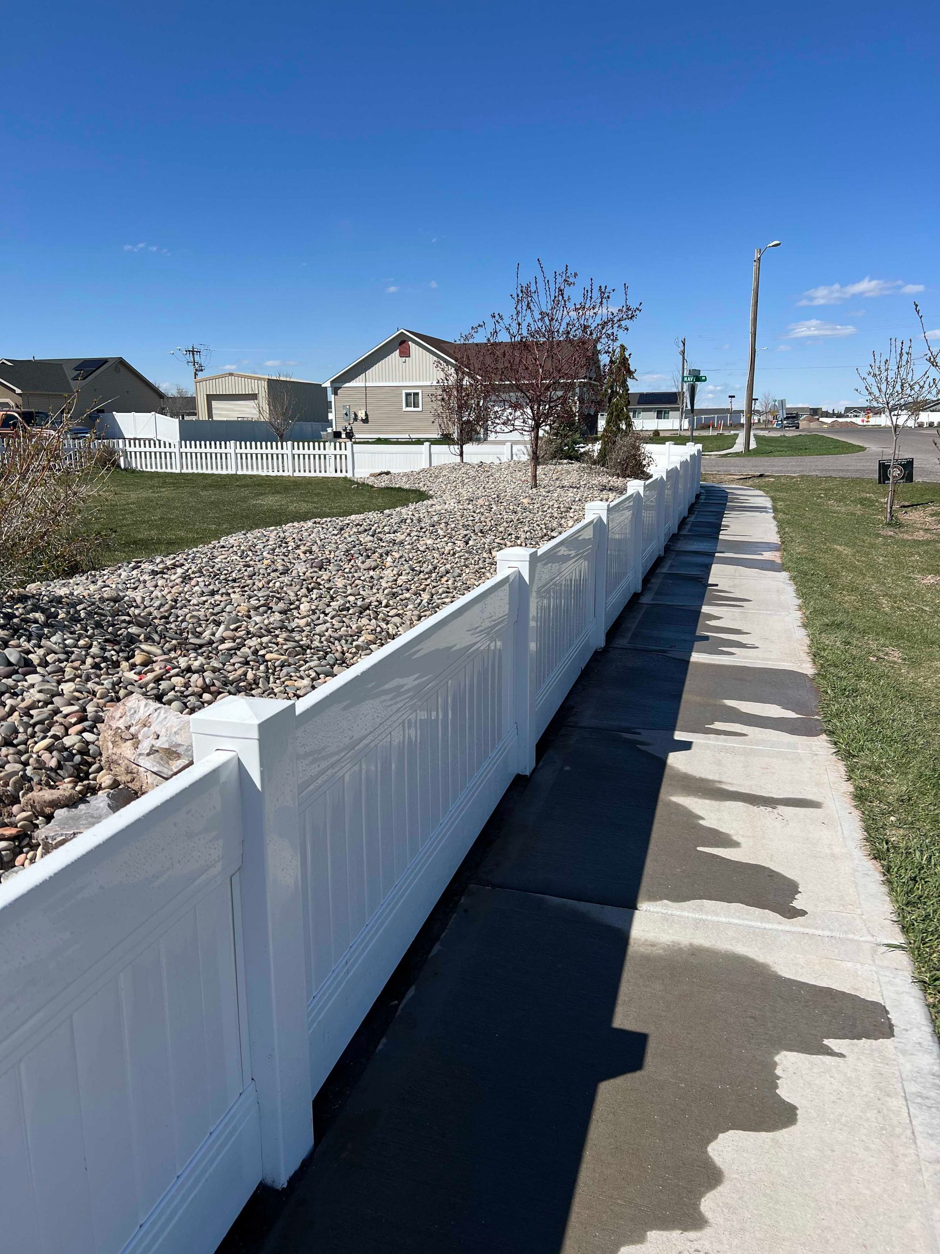 White picket fence along a sidewalk, bordering a rock-filled landscape. Bright blue sky.