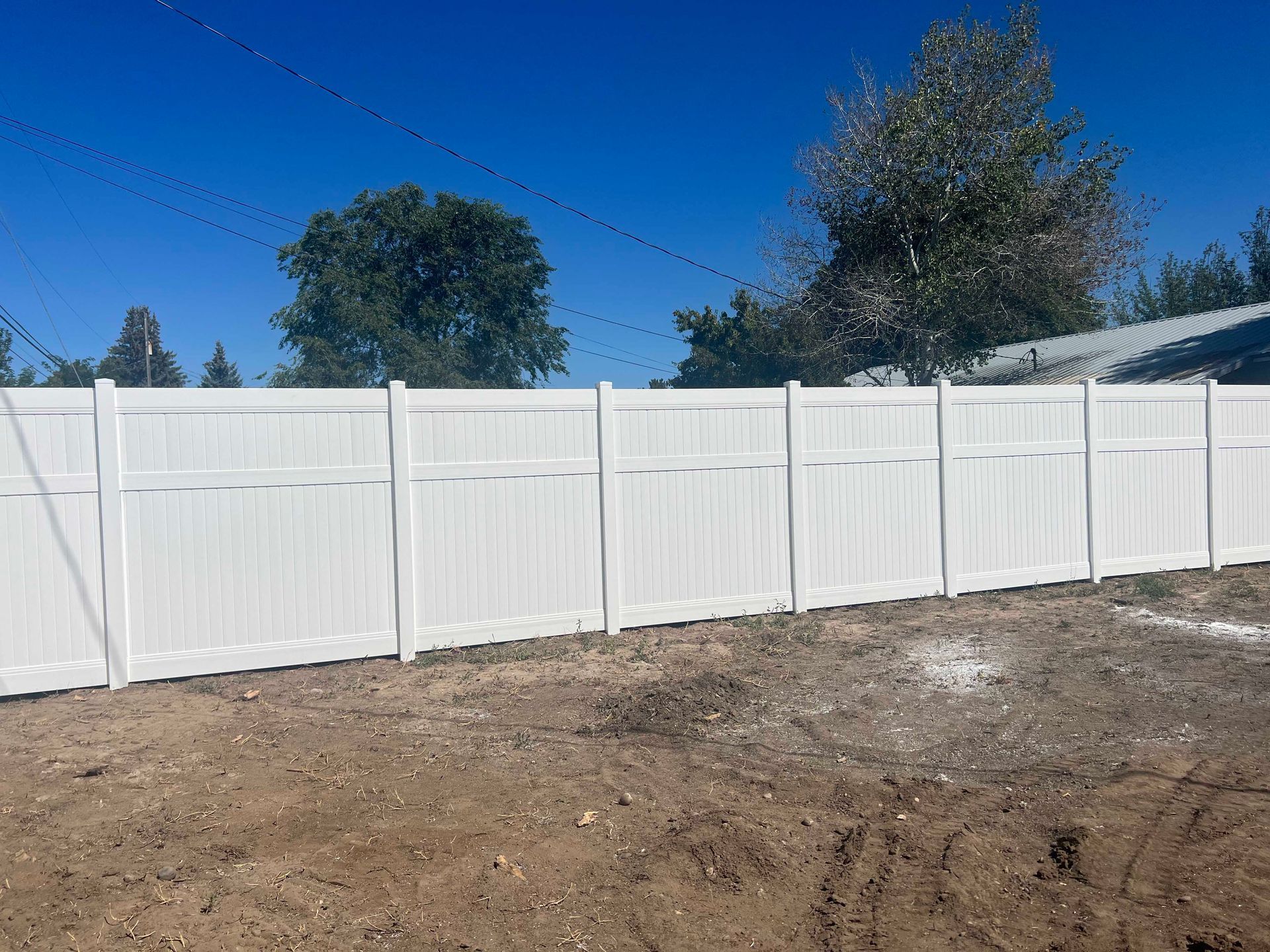 White vinyl fence against a blue sky, enclosing a dirt area with trees in the background.