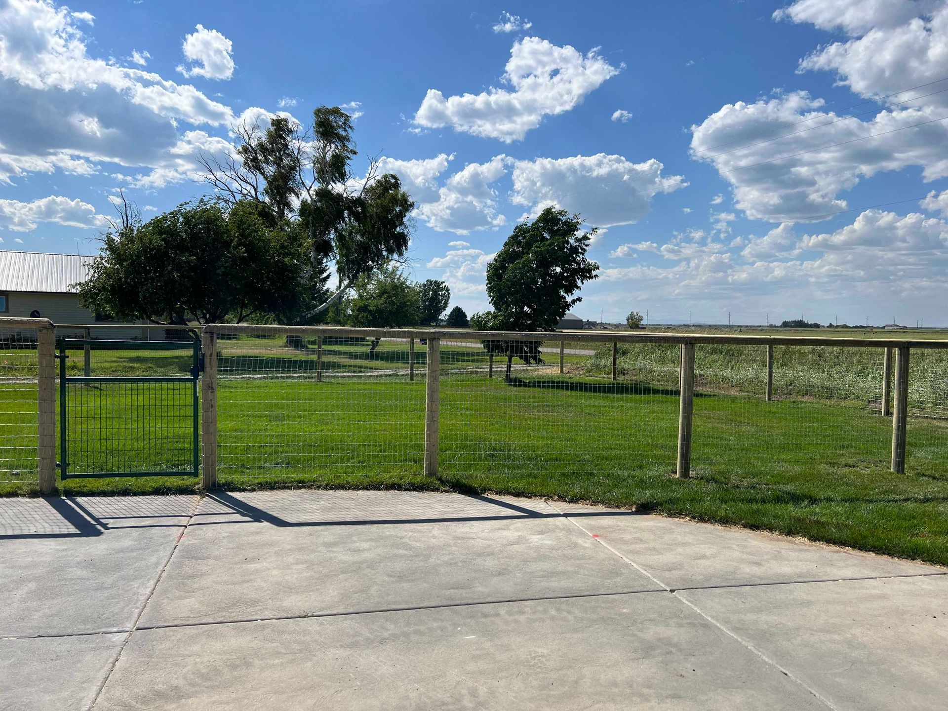 Green fenced yard, concrete patio, trees, and cloudy blue sky.