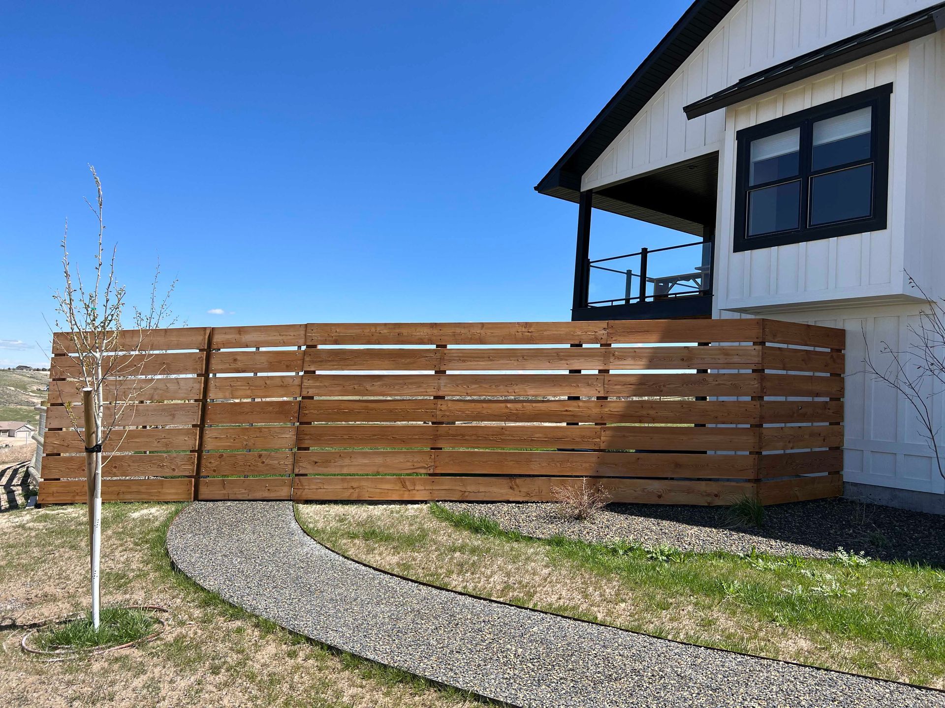 Wooden fence in front of a white house with a black-framed window, gravel path in the foreground under a blue sky.