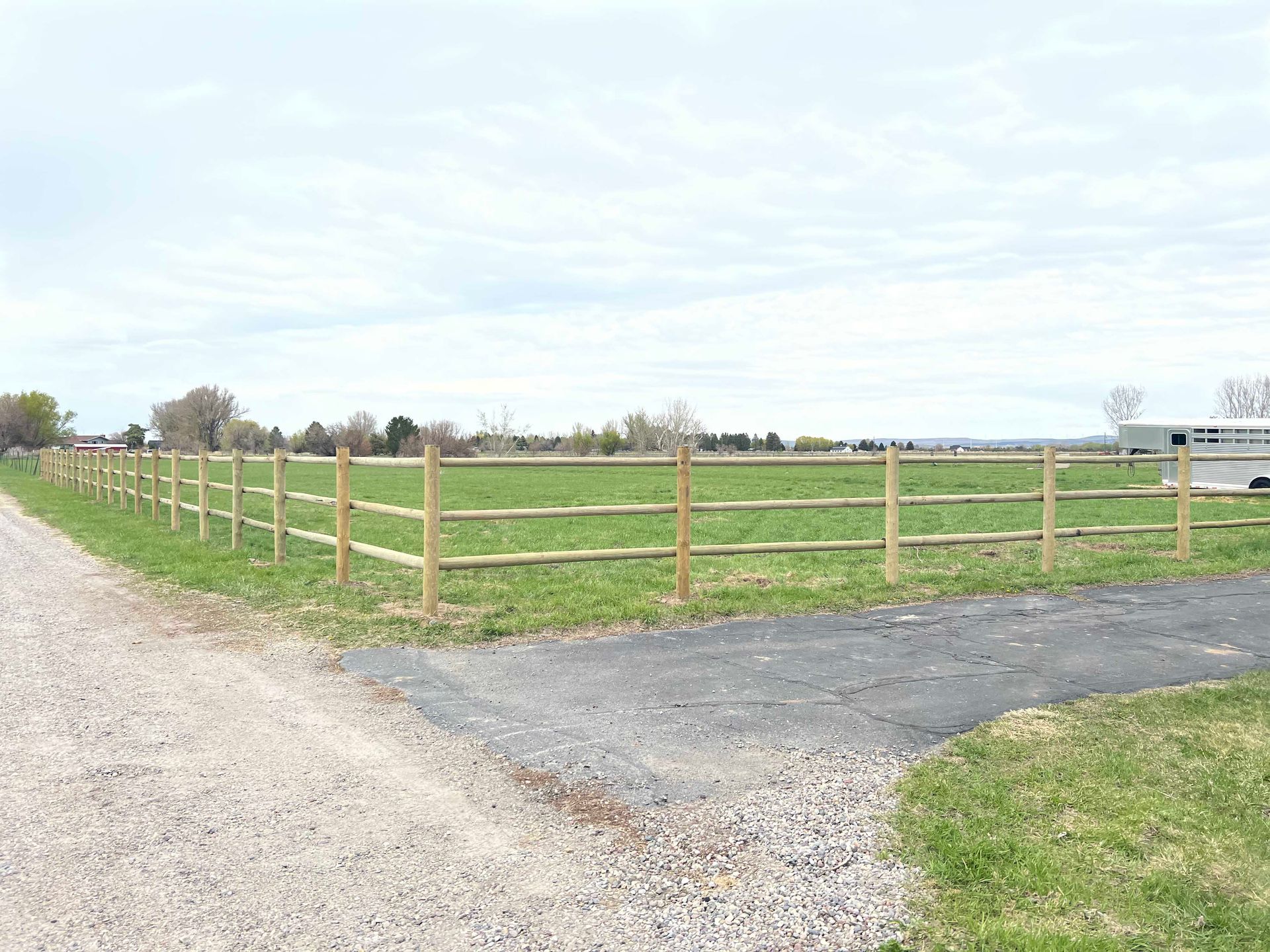 Wooden split-rail fence surrounds a grassy field, next to a gravel driveway under a cloudy sky.
