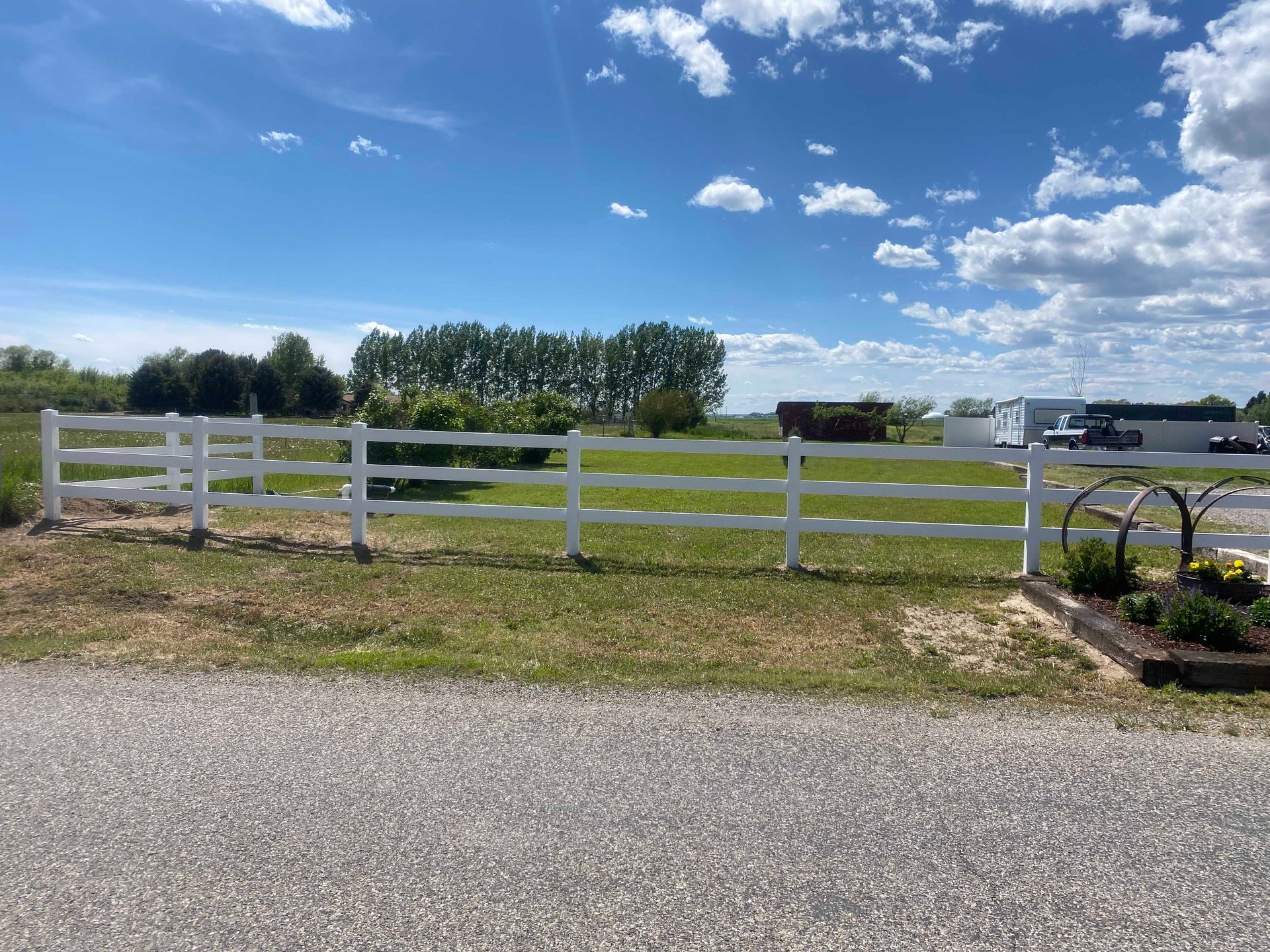 White fence in a grassy field under a blue sky with fluffy clouds. Trees and buildings in the background.
