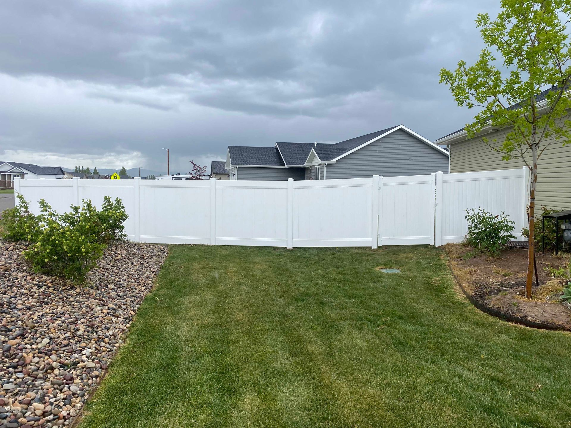 White vinyl fence in a backyard, with green grass and a house in the background under a cloudy sky.