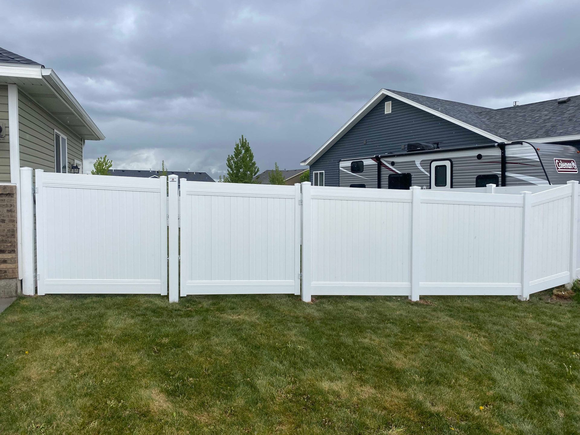 White vinyl fence encloses a green lawn, near a house and RV, under a cloudy sky.