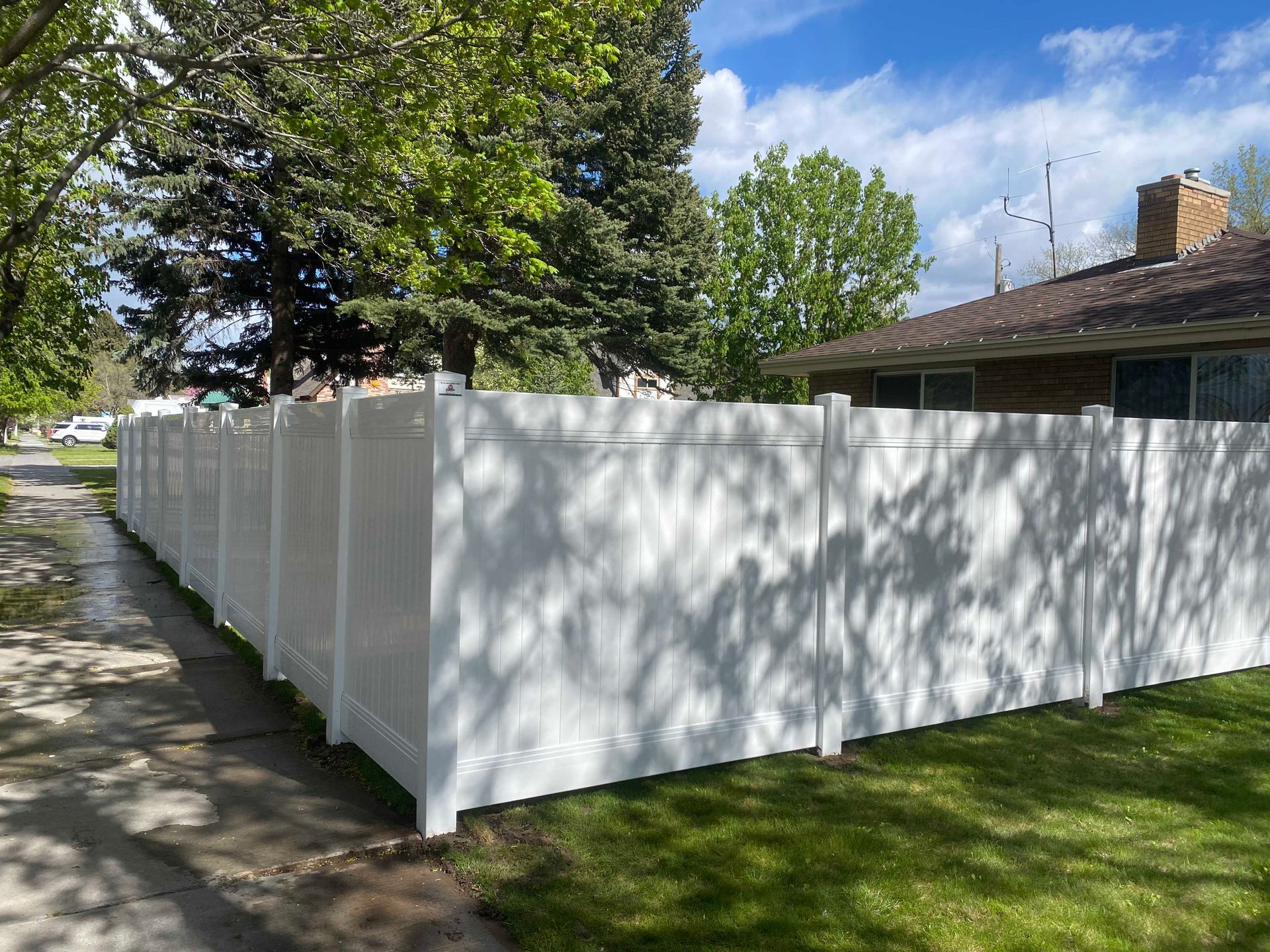 White vinyl fence along a sidewalk and grass lawn, adjacent to a house with a brick chimney under a blue sky.