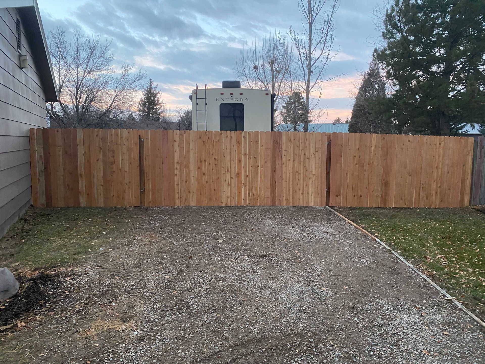 Wooden privacy fence in front of a gravel driveway, with RV and trees visible beyond it, at dusk.