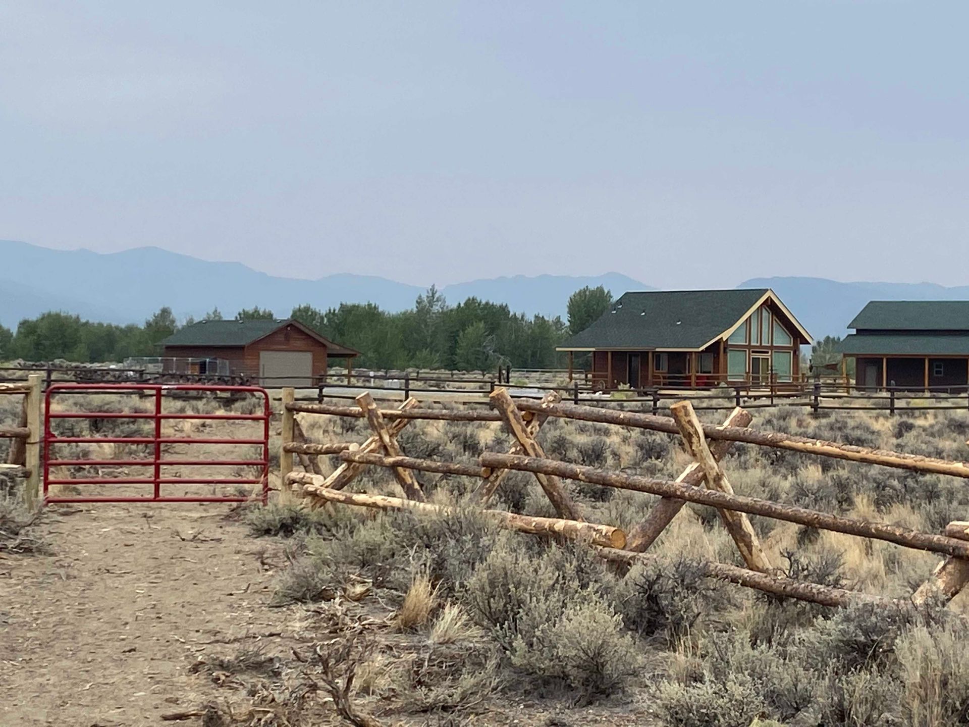 Ranch with log fence, buildings, and gate, with mountains in the background under a hazy sky.