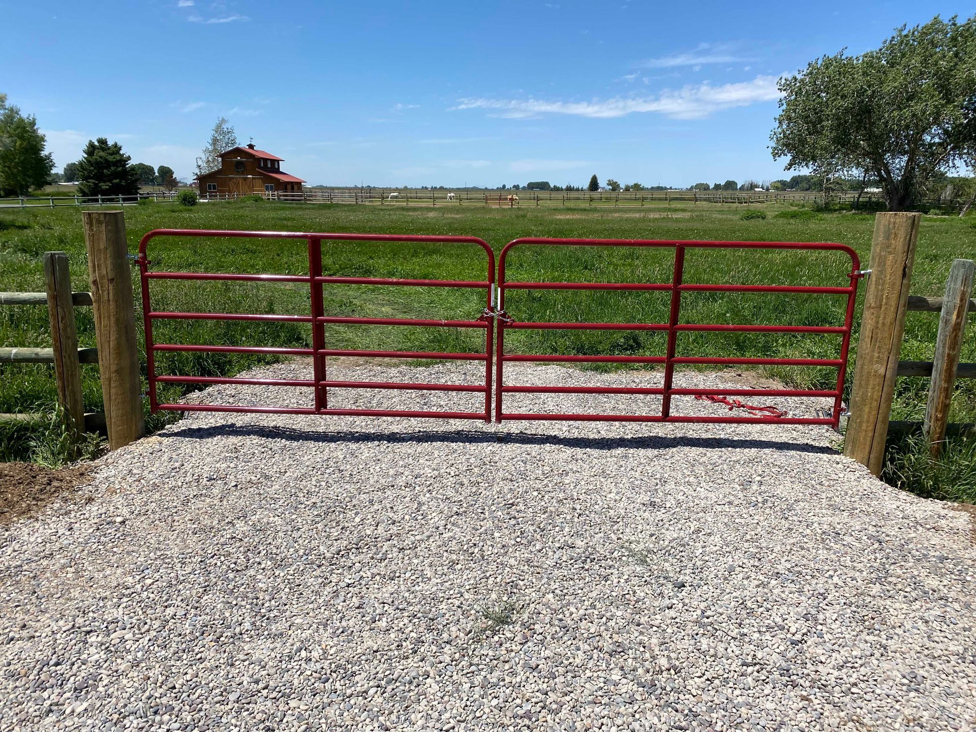 Red metal farm gate on gravel, leading to a field with a barn in the distance under a blue sky.