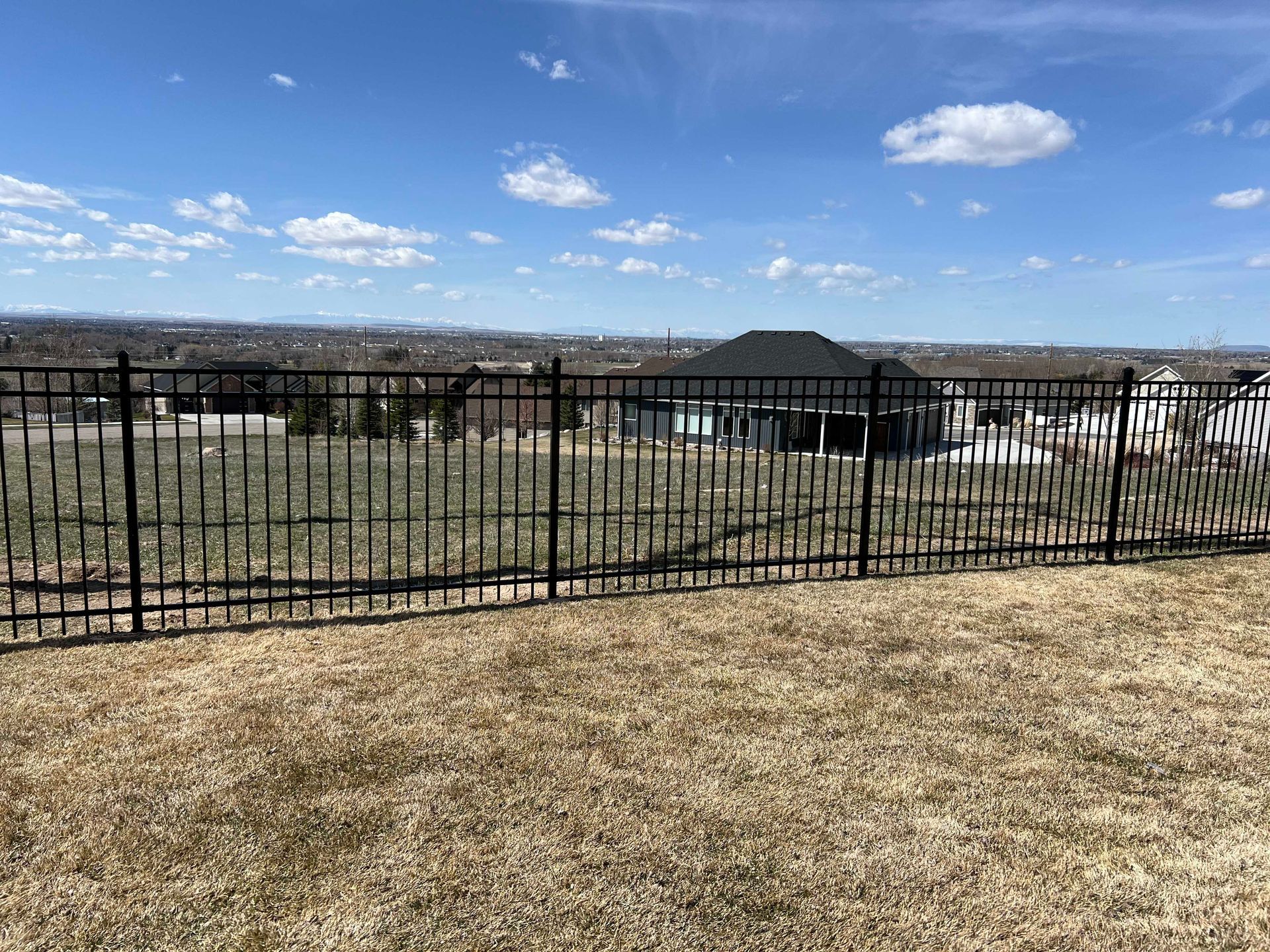 Black metal fence on a grassy hill overlooking a suburban landscape under a blue sky with clouds.