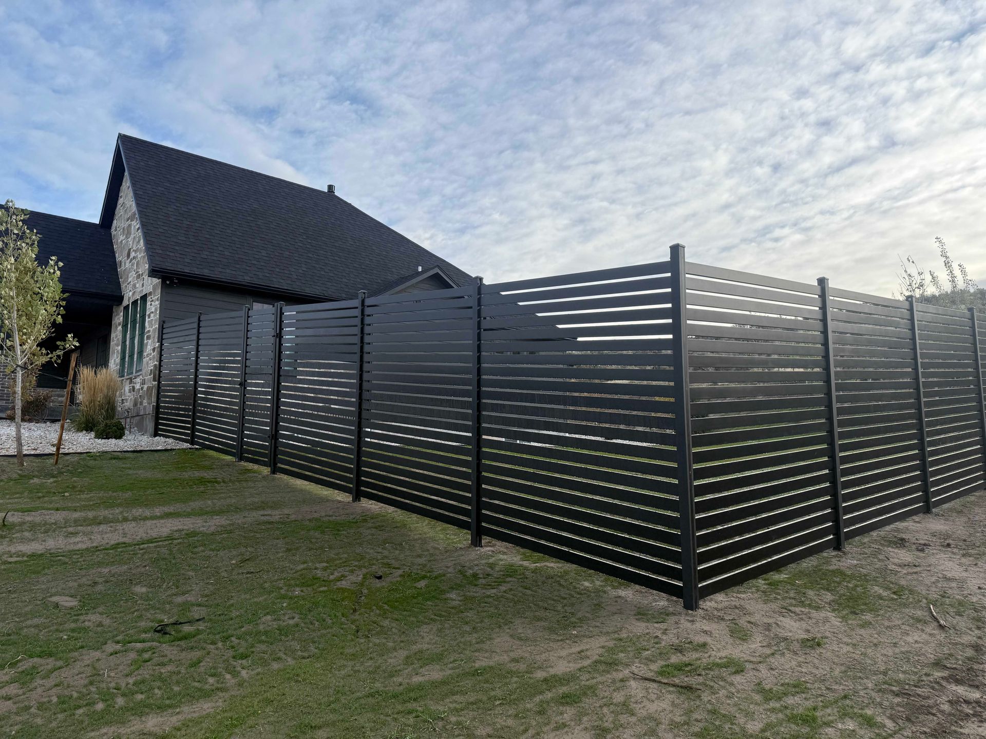 Black horizontal slat fence surrounds a house with a dark roof on a cloudy day.