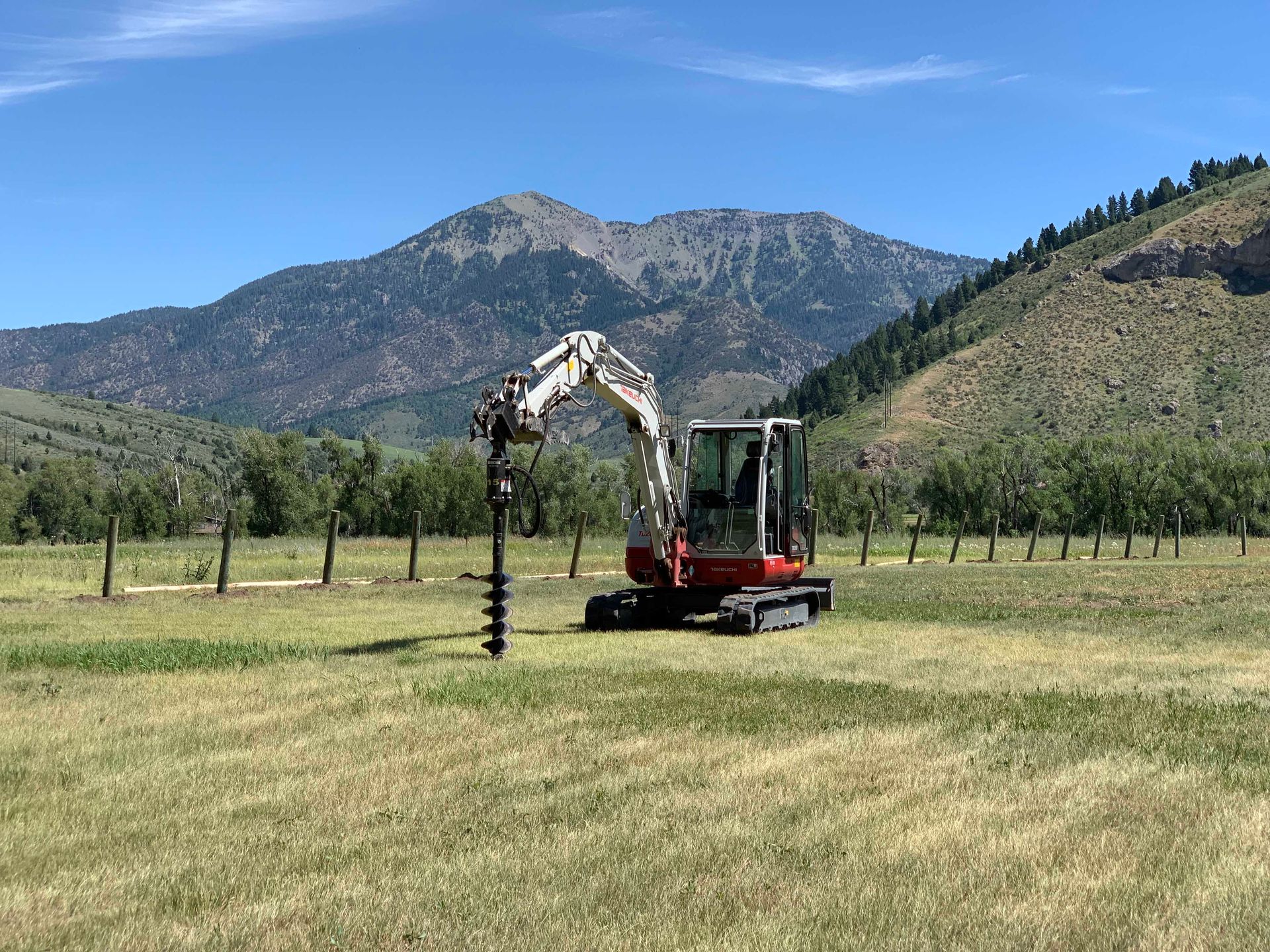 Excavator drilling holes in a field for fence posts, mountain backdrop.