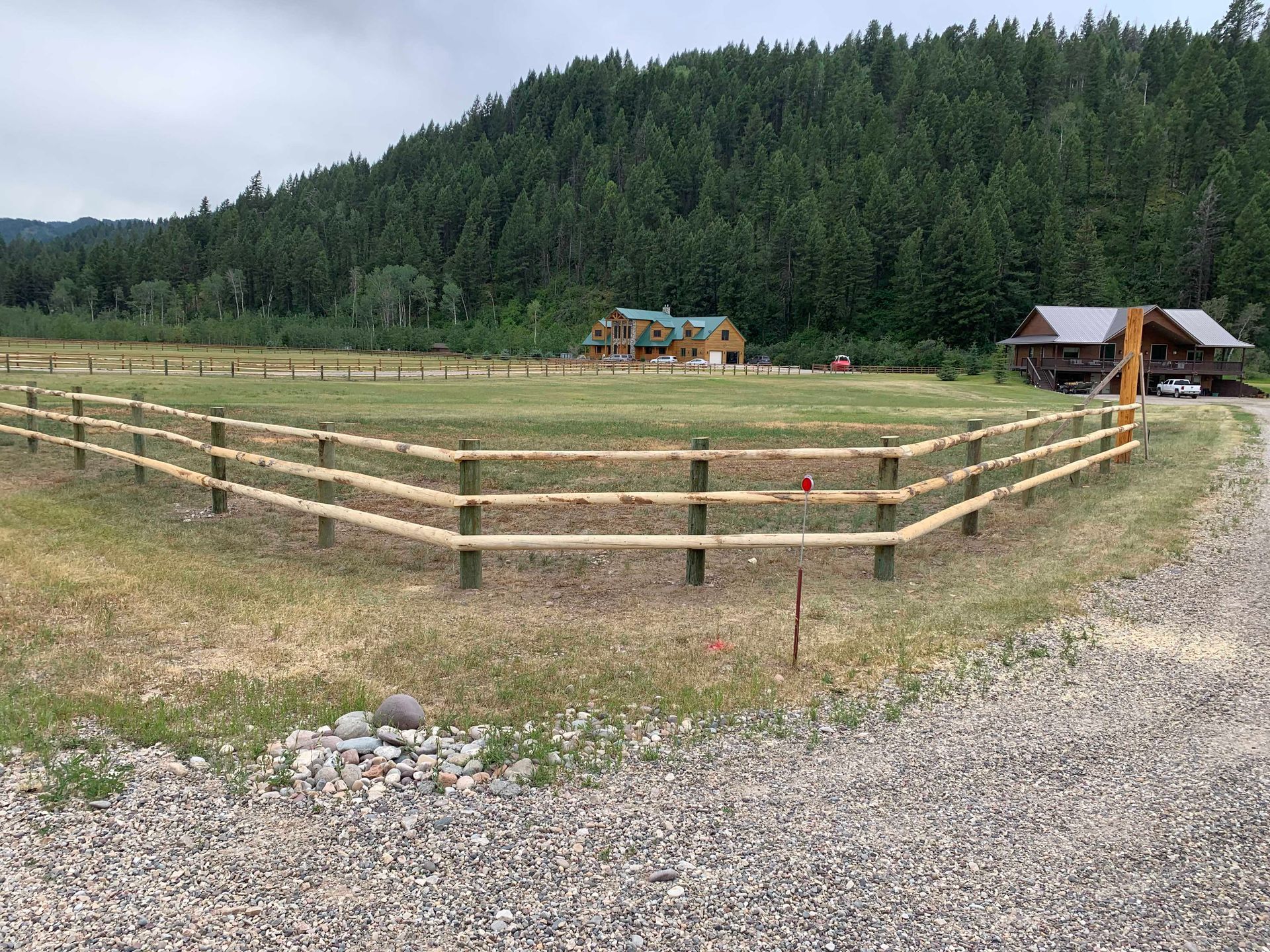 A wooden split-rail fence surrounds a grassy field with two cabins and a forest backdrop.