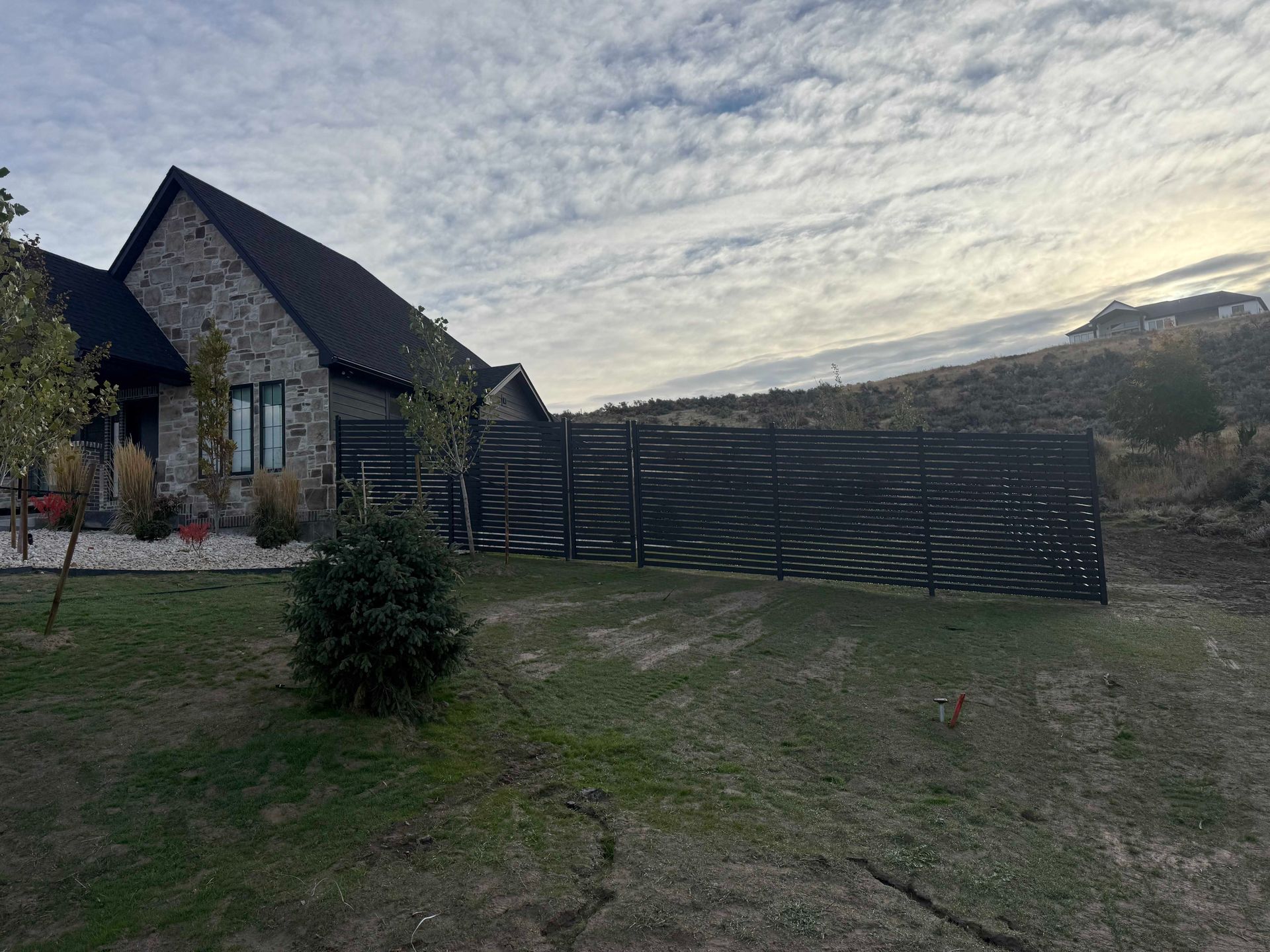House with stone facade and black fence on a grassy hill, under a cloudy sky.
