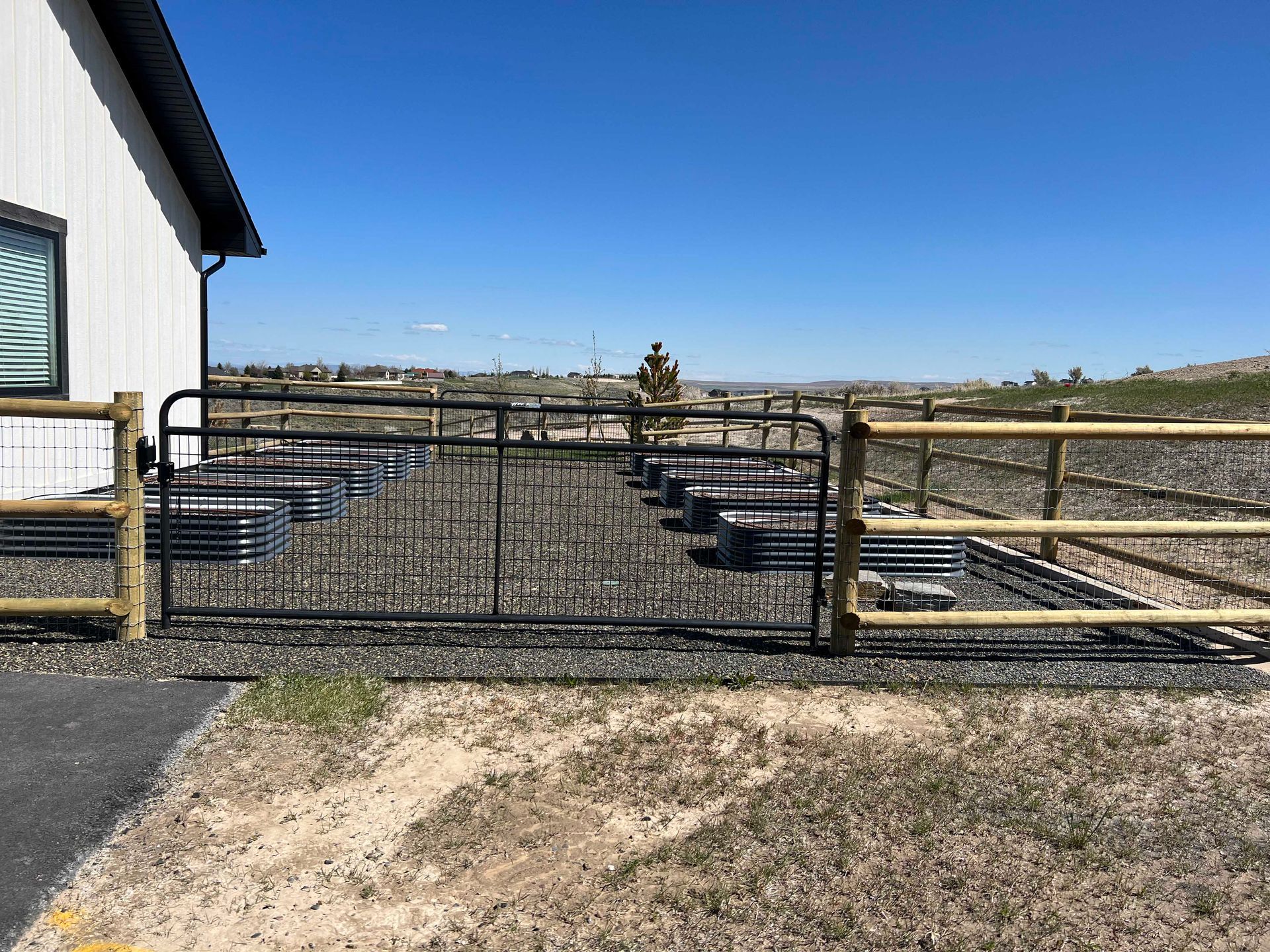 Black metal gate in front of raised garden beds. Brown wooden fence sections on either side, house in the background.