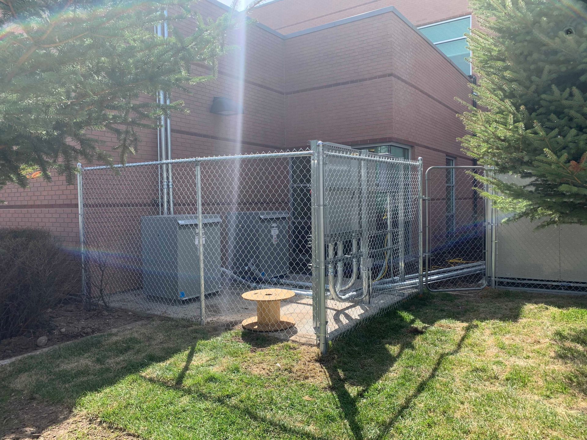 Chain-link fenced enclosure with electrical equipment next to a brick building.