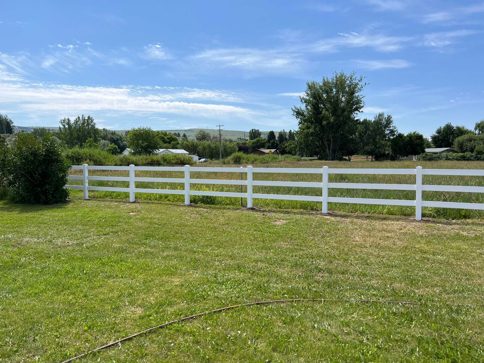 White fence in a grassy field on a sunny day. Background includes trees, buildings, and a blue sky.