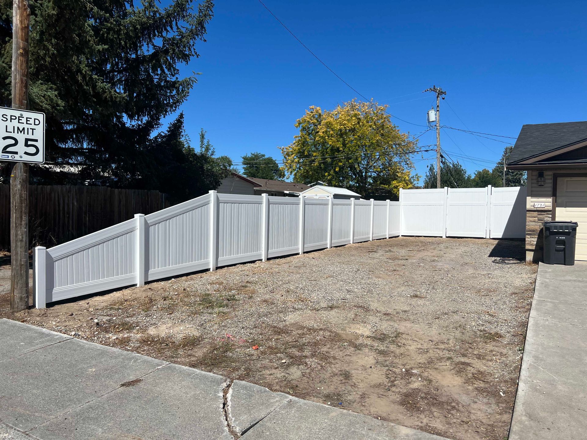 White vinyl fence surrounds a gravel yard on a sunny day next to a house. Speed limit sign.