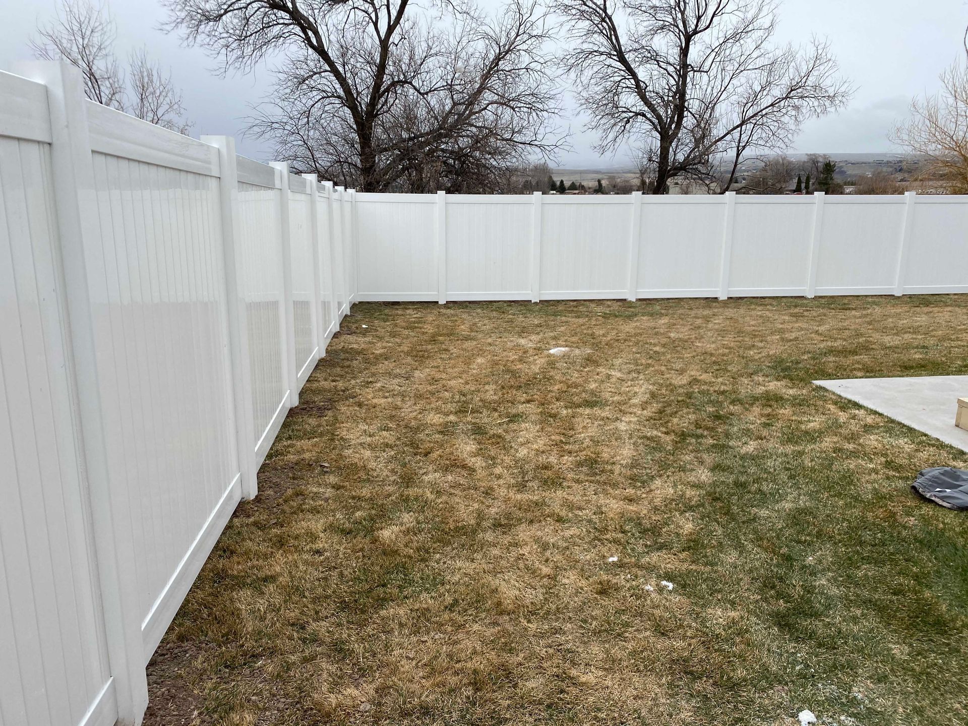 White vinyl fence enclosing a brown, patchy lawn on an overcast day. Bare trees and distant hills are visible.