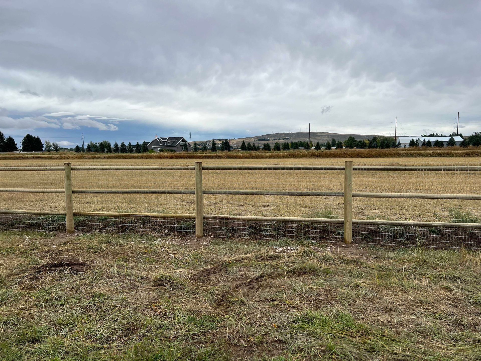 Wooden fence in a field with dry grass, with a town and cloudy sky in the background.