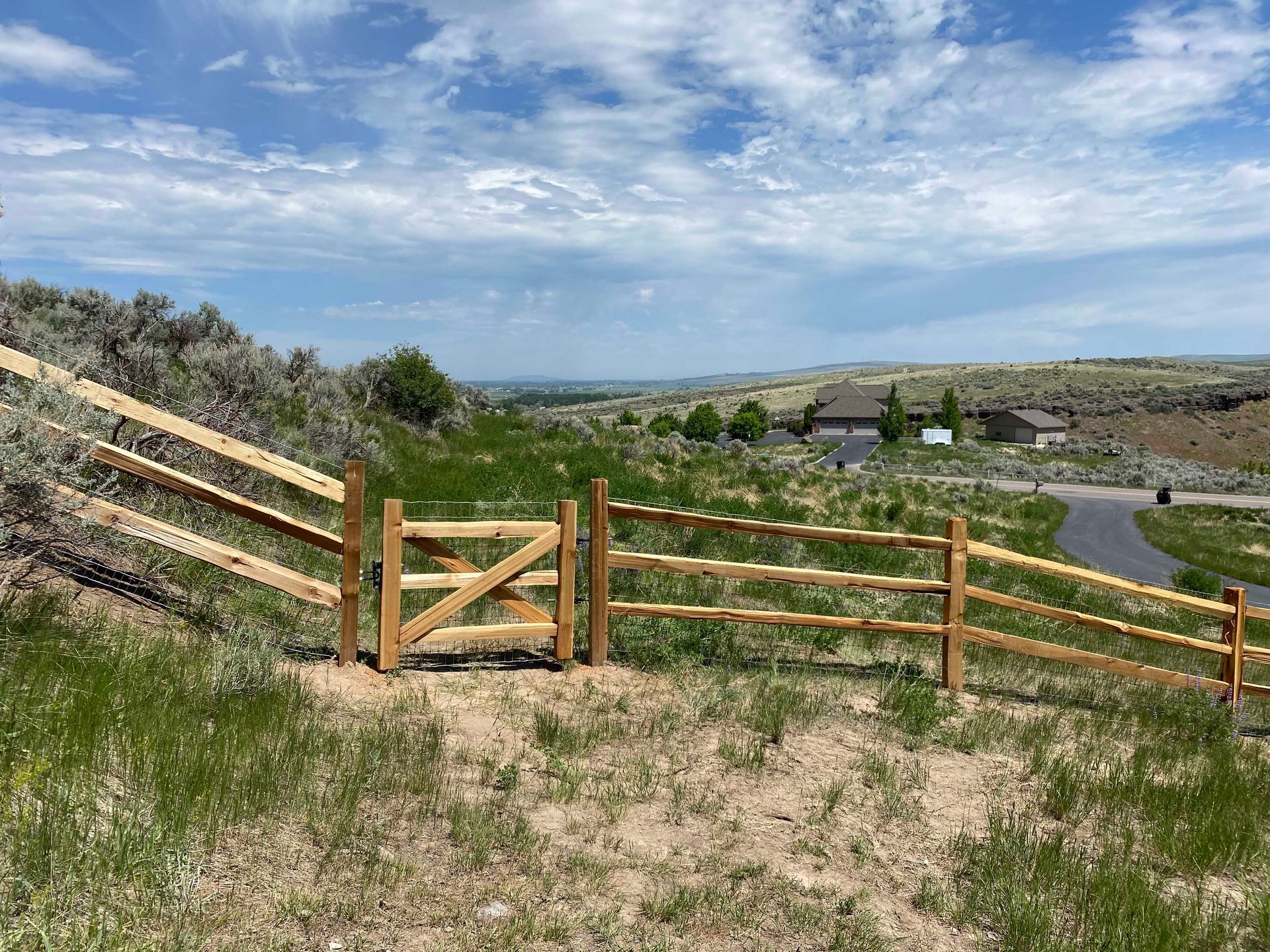Wooden split-rail fence with gate on a grassy hillside, overlooking a distant residential area under a cloudy sky.