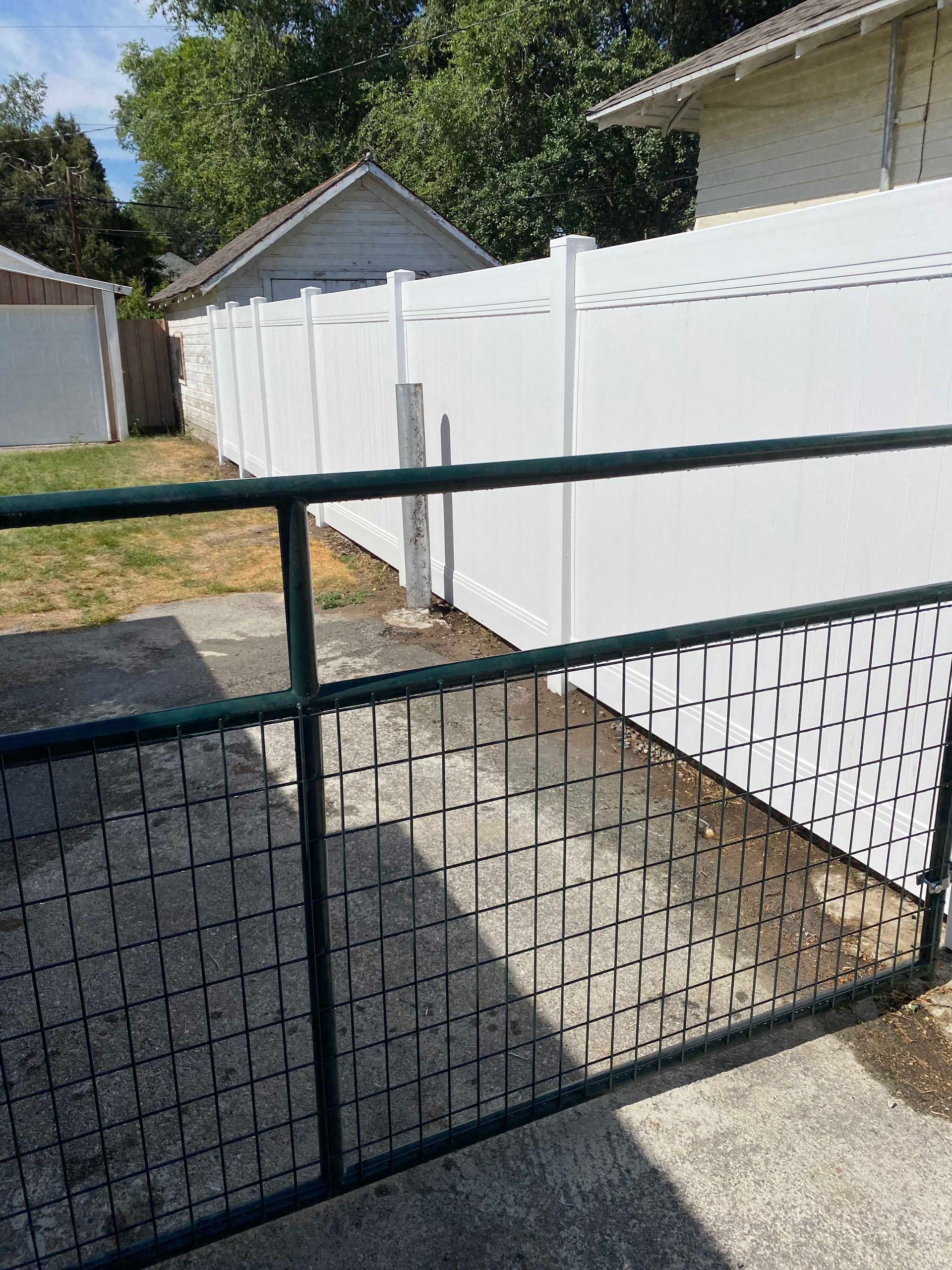 Green metal gate leading to a white vinyl fence in a yard with a concrete surface.