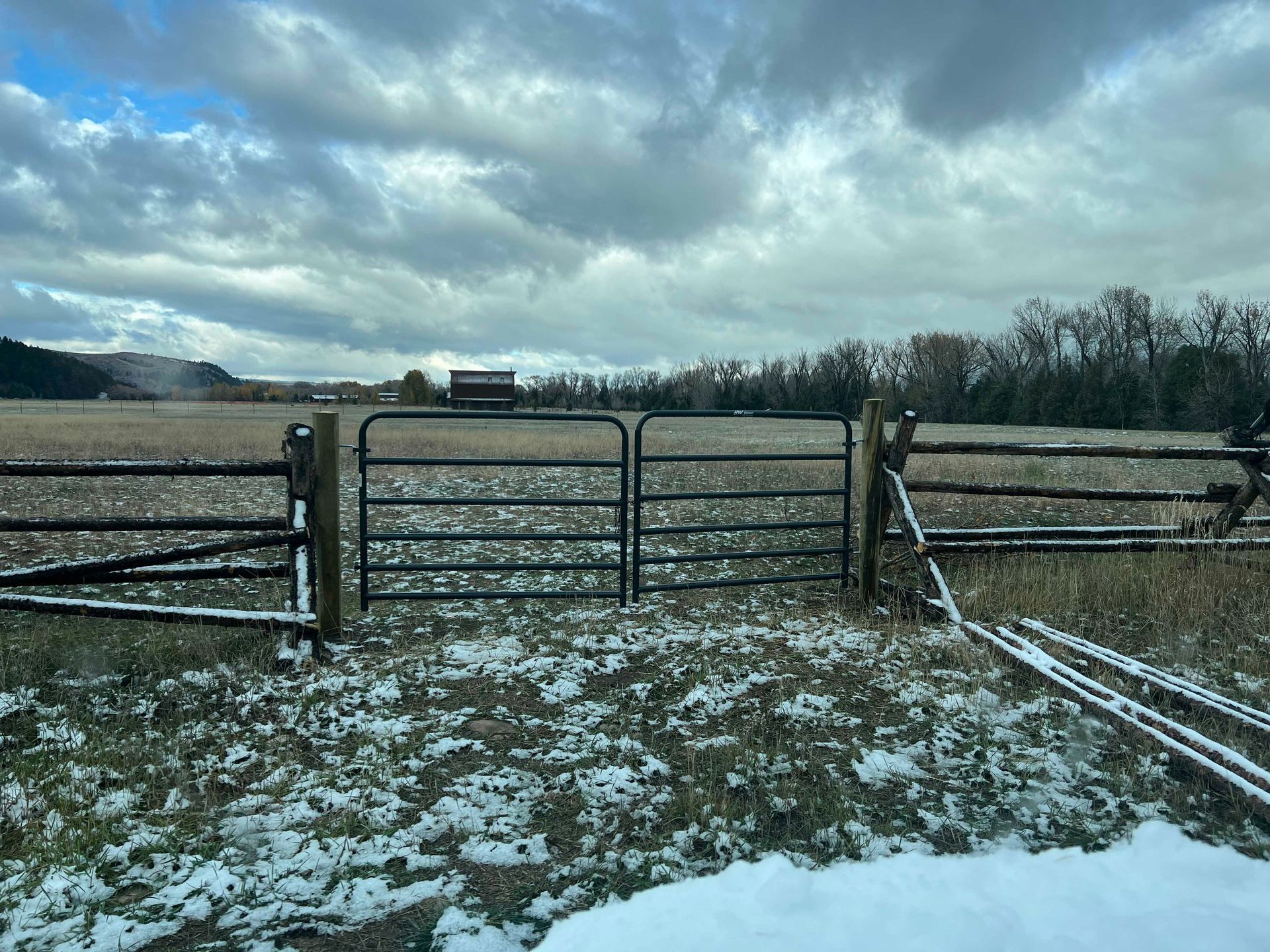 Snow-dusted field with a closed black metal gate. Cloudy sky over the rural landscape.