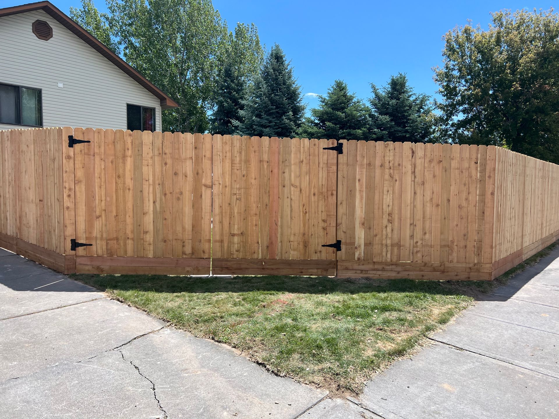 Wooden fence with black hinges and a house in the background. Grass and sidewalk in front.