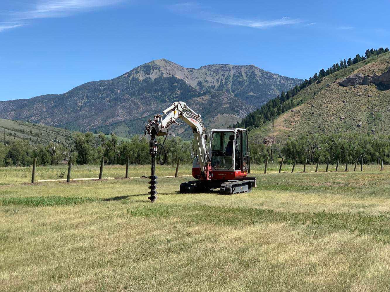 Excavator with auger drilling in a field, mountains in background, sunny day.