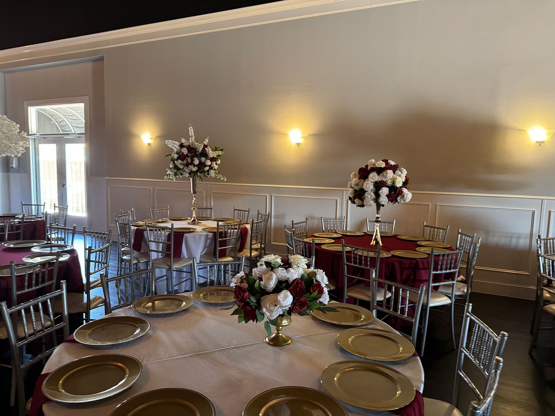Elegant banquet hall set for dining; gold chargers, floral centerpieces, maroon table linens.