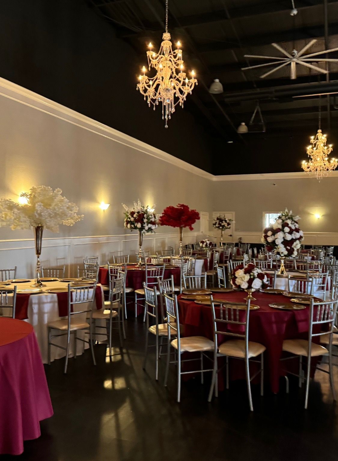 Event hall with tables set for a banquet; maroon tablecloths, gold chairs, and floral centerpieces; crystal chandeliers.