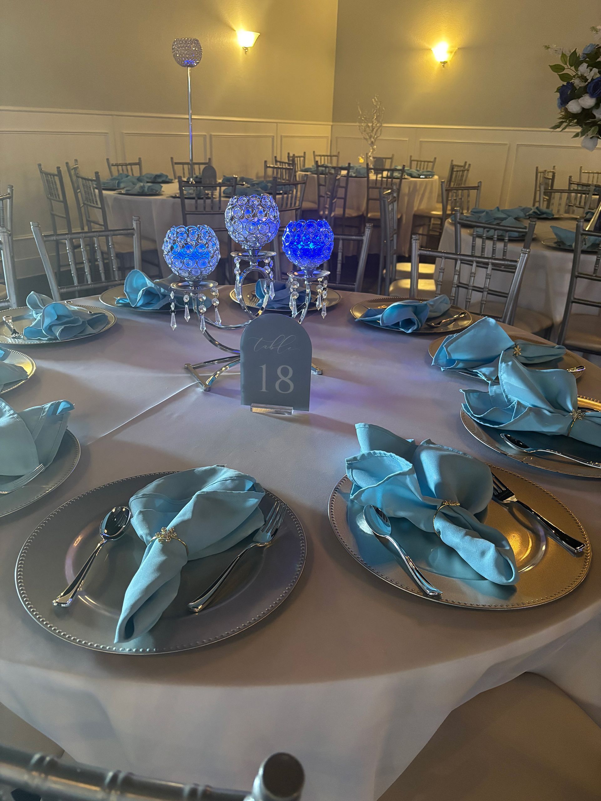 Round table set for a formal event, featuring silver chargers, light blue napkins, and centerpieces with blue lights.