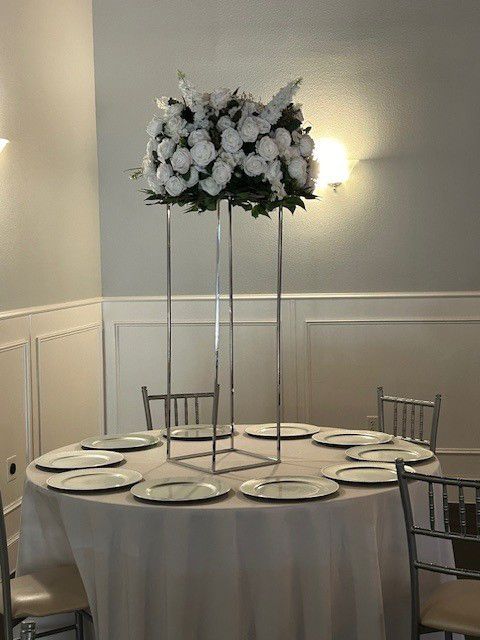 White floral centerpiece on tall silver stand atop round table set for a meal.