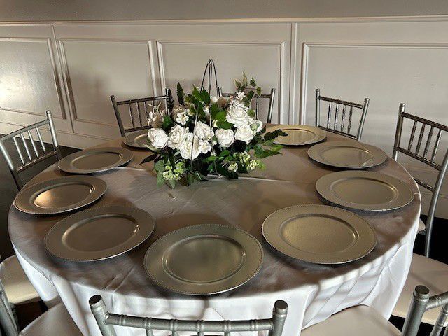 Round table set for a formal event with white flowers, silver charger plates, and silver chairs.