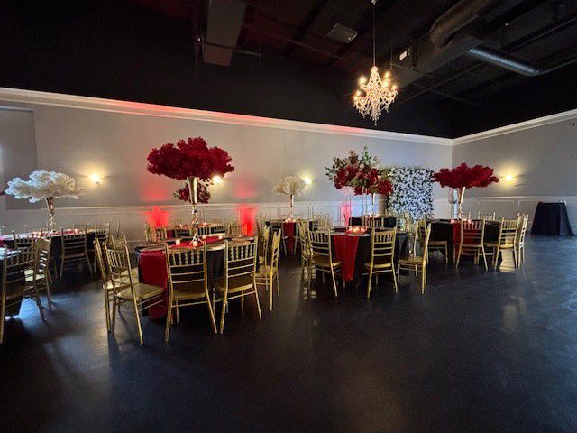 Elegant event space with round tables, red and black linens, gold chairs, and floral centerpieces.