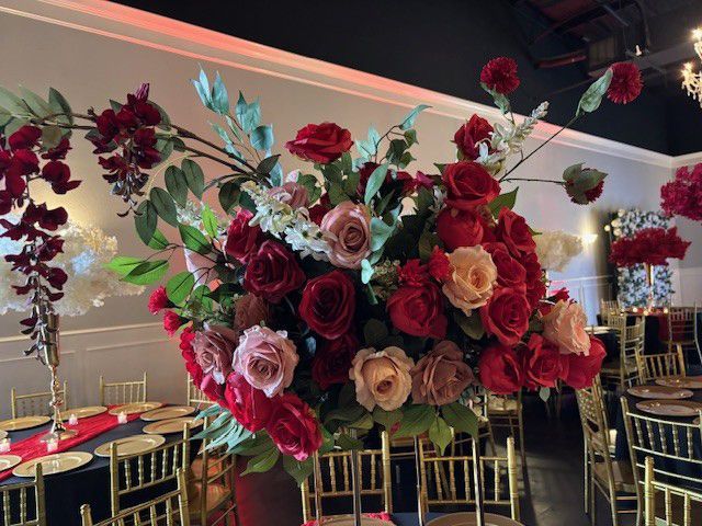 A large floral arrangement with red and pink roses, set in a dining room with gold chairs.
