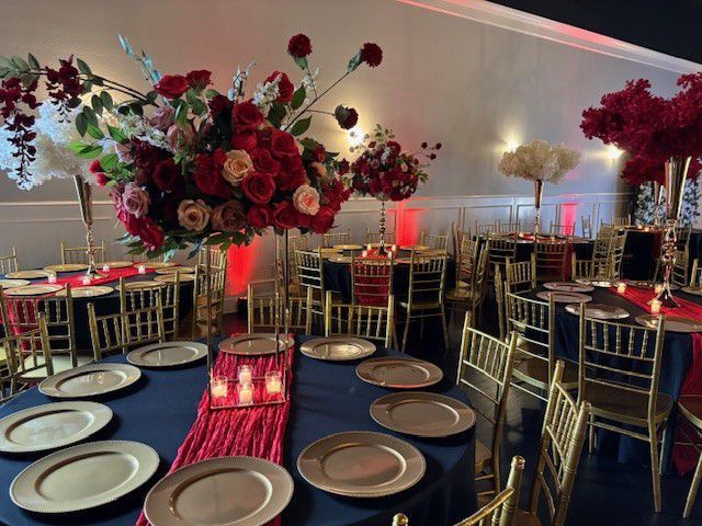 Elegant event hall with round tables, gold chairs, navy tablecloths, red accents, and large floral centerpieces.