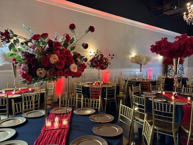 Elegant event space with round tables, floral centerpieces in red and gold, and gold chairs.