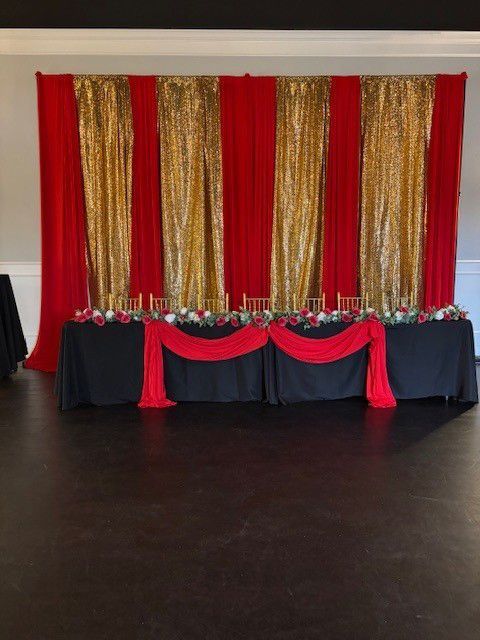 Red and gold event backdrop behind a black draped table with red and white floral arrangements.