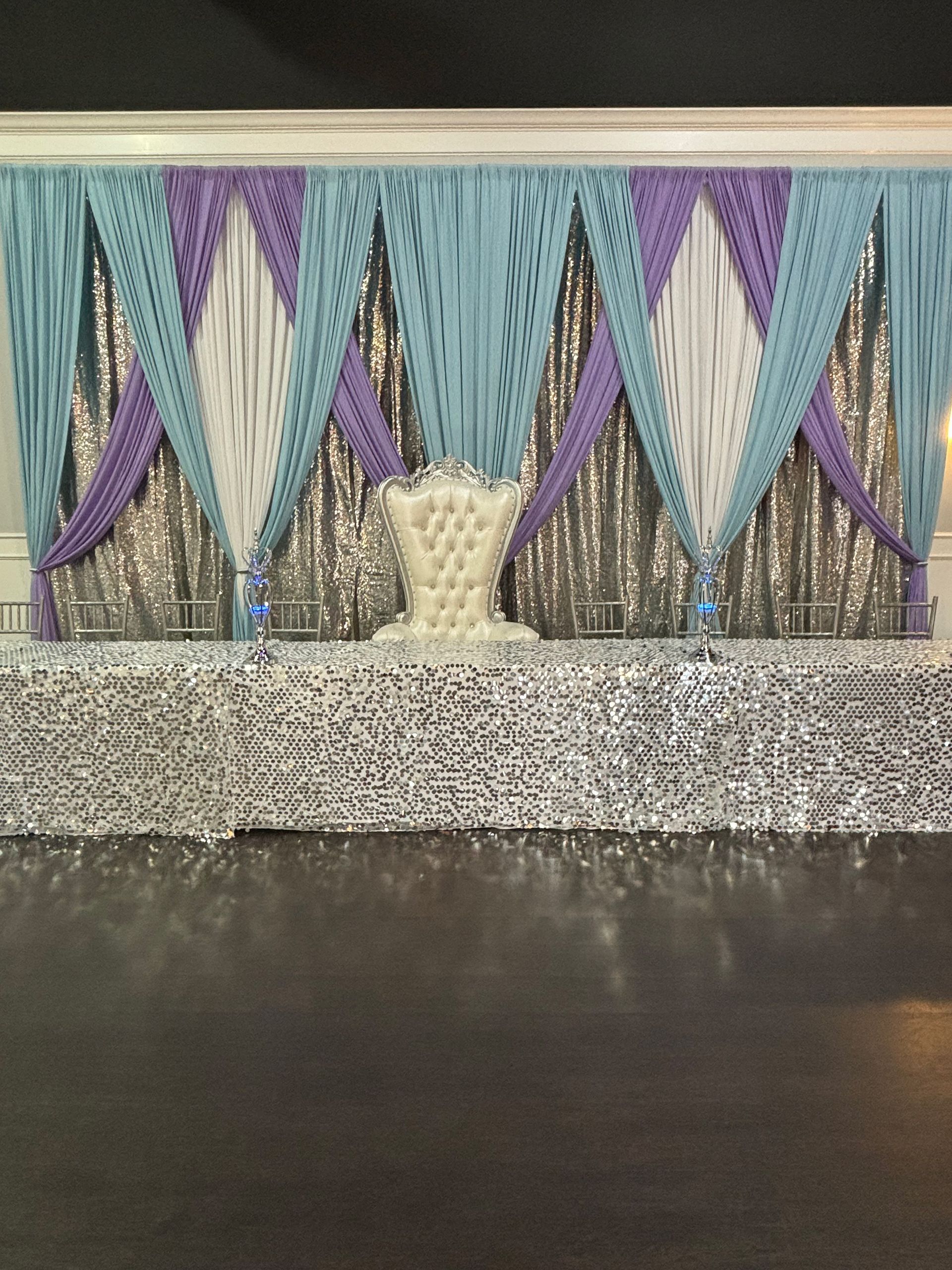 Stage with sequin backdrop, draped fabric in blue, purple, white. White throne chair centered.