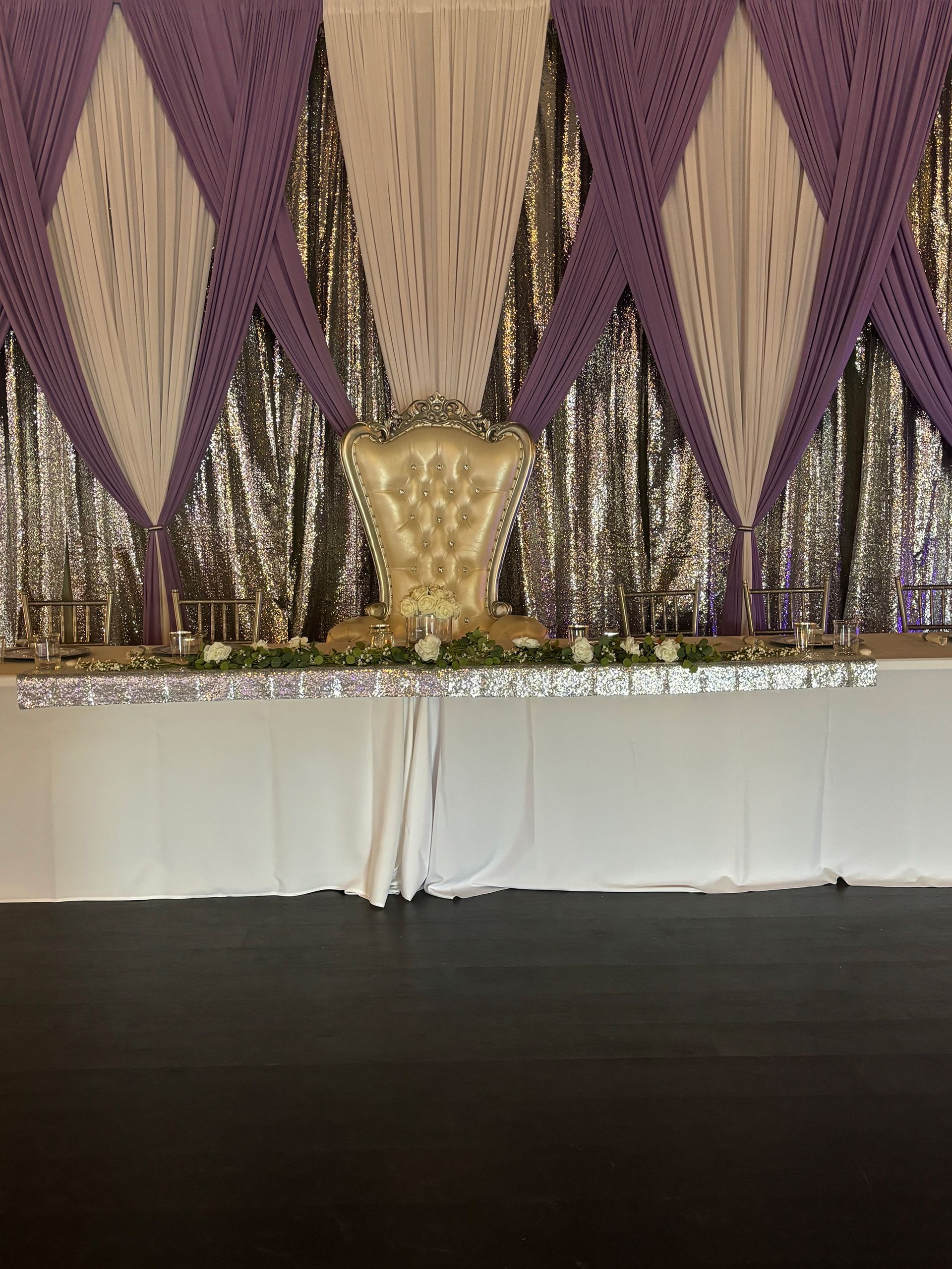 Purple and silver decorated event backdrop with a throne-like chair centered on a white table.