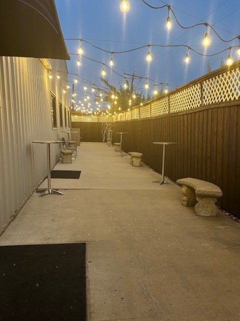 Outdoor patio with string lights, concrete floor, tables, benches, and a wooden fence.