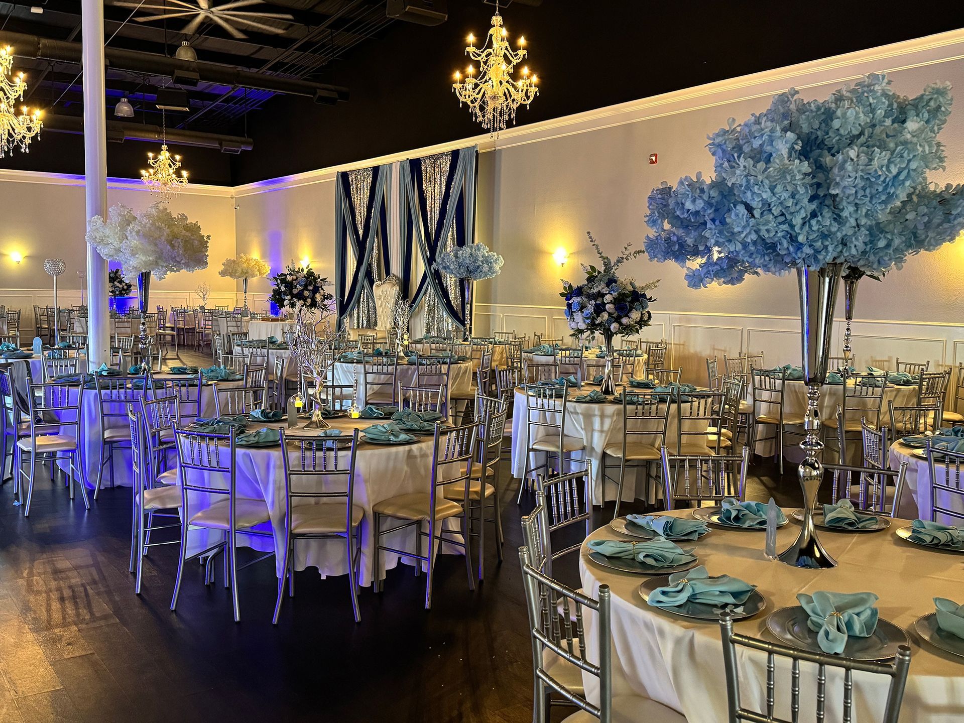 Elegant event hall with round tables set for a celebration; blue and silver decor, chandeliers, and floral centerpieces.