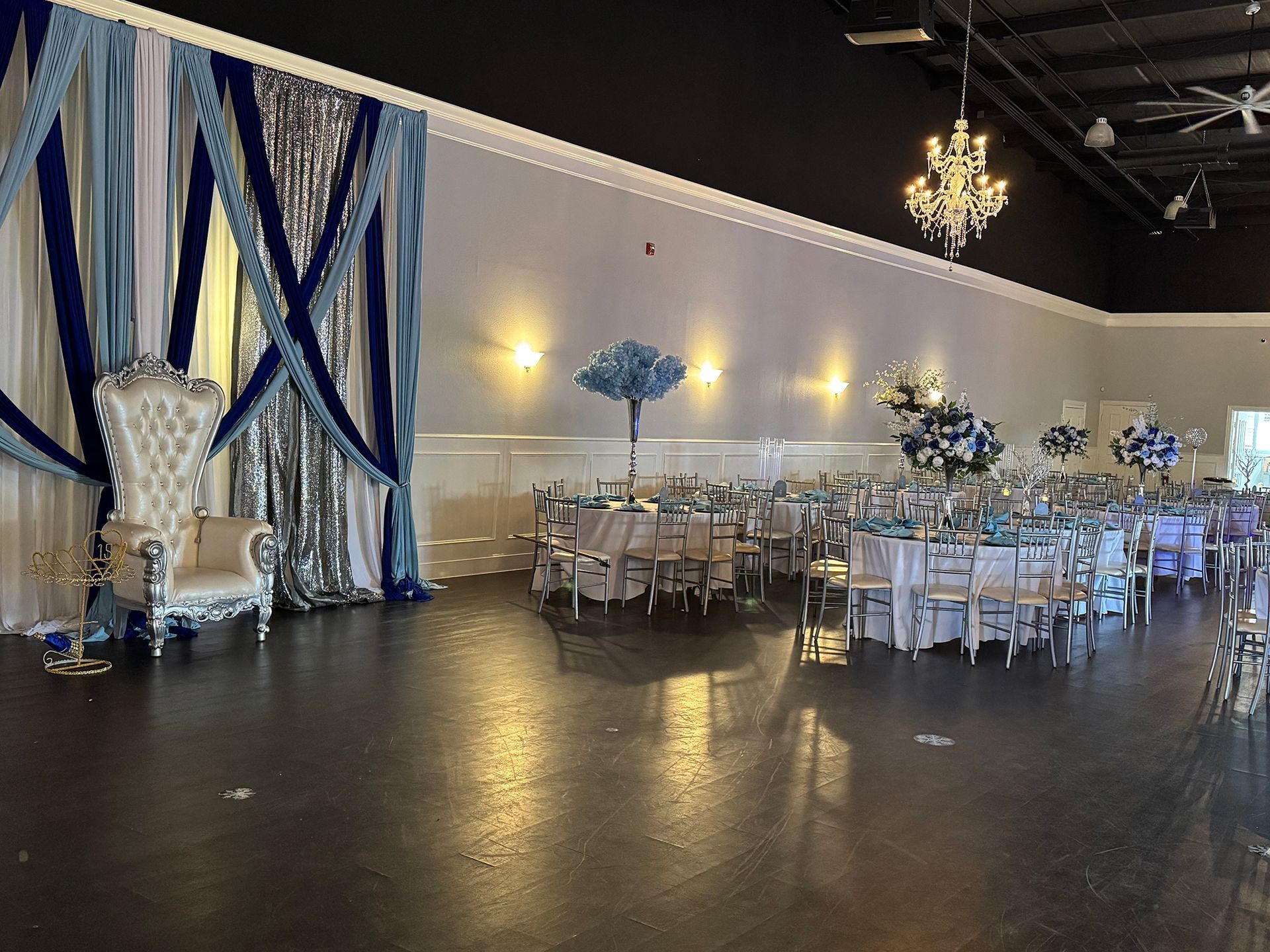 Elegant event hall with blue, silver decor; throne chair, tables, chandeliers, dark floor.