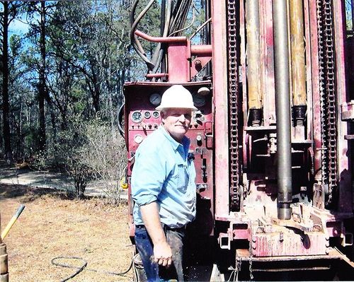 Worker in a white hard hat stands beside a red drilling rig in a wooded outdoor area.
