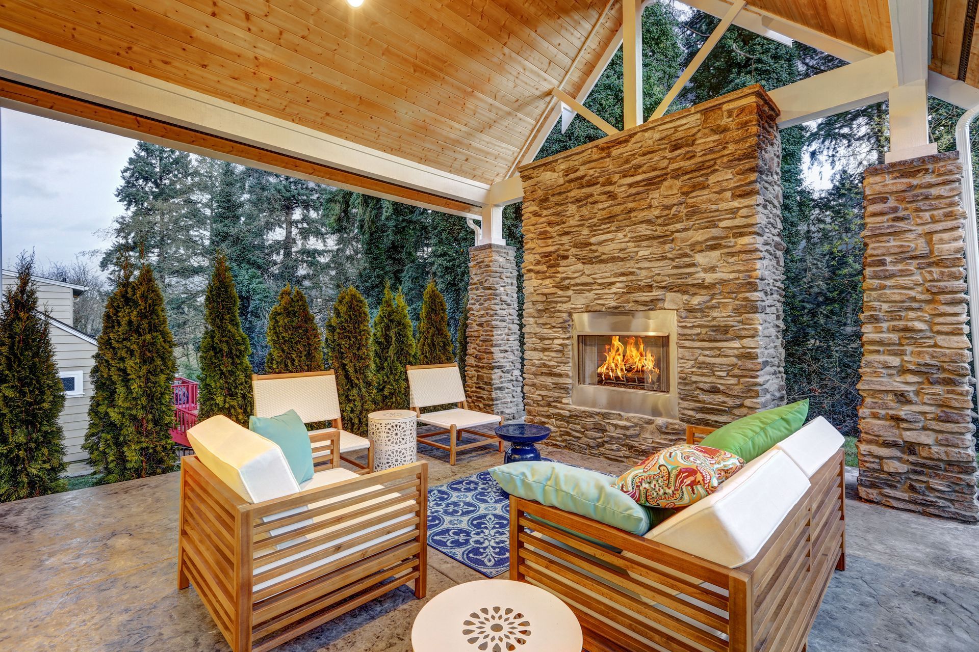 Outdoor patio with fireplace, seating, and wooden beams, overlooking a green space.