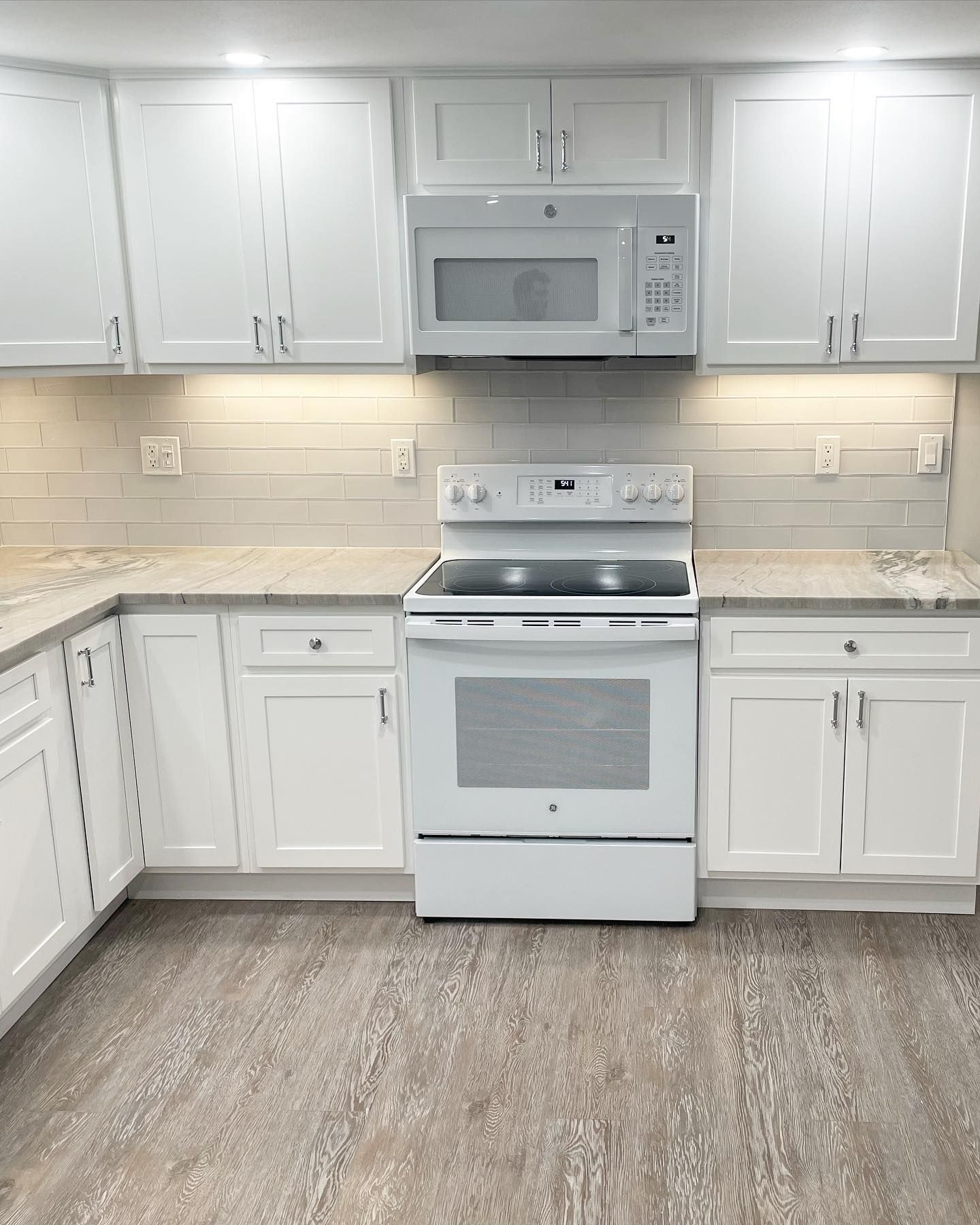 White kitchen with cabinets, stove, microwave, light gray backsplash, and wood-look flooring.