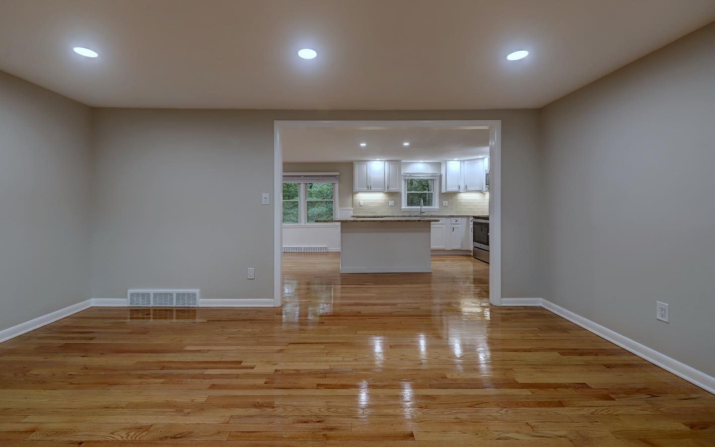 Empty room with hardwood floors, a doorway leading to a kitchen with white cabinets, and recessed lighting.